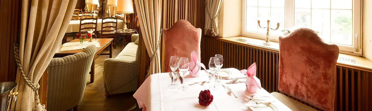 Tischgedeck in einem eleganten Restaurant mit rosa Tischdecke, Gläsern, Besteck und einer roten Rose als Tischdekoration. Im Hintergrund sind Stühle und Vorhänge sichtbar.