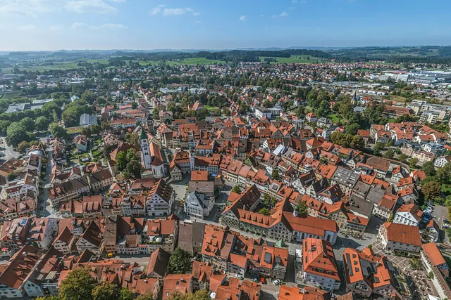 Luftaufnahme einer Stadt mit roten Dächern, umgeben von grünen Feldern und Hügeln. Sichtbare Gebäude in verschiedenen Formen und Größen.