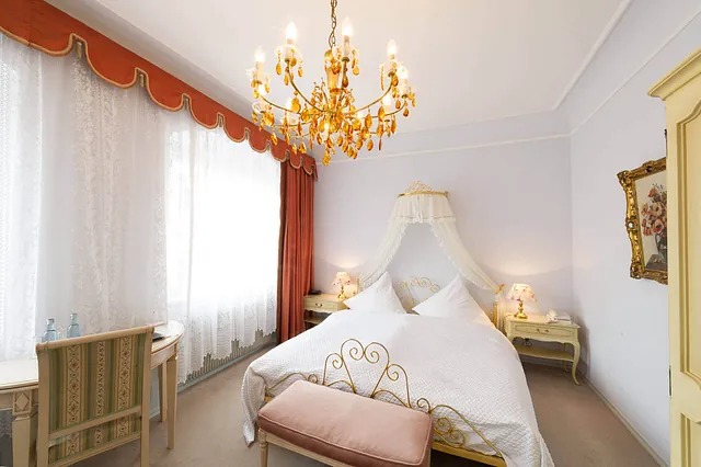 Schlafzimmer mit einem großen Bett, einem Kronleuchter und einem gepolsterten Hocker. Vorhänge in Orange und weiße Gardinen sind sichtbar.