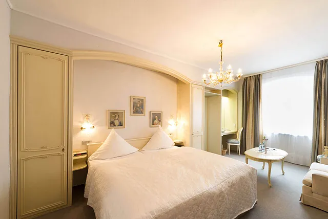 Helles Hotelzimmer mit einem großen, weißen Bett, zwei Kissen in Form von Spitzen, einem Kronleuchter, zwei Wandlampen und Bildern an der Wand.