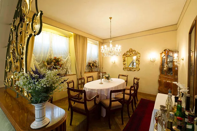 Ein eleganter Restaurantraum mit einem runden Tisch, umgeben von sechs Stühlen. An der Wand hängen zwei Spiegel und eine goldene Wandverzierung. Ein Kronleuchter hängt von der Decke. Auf dem Tisch steht eine Blumenvase.