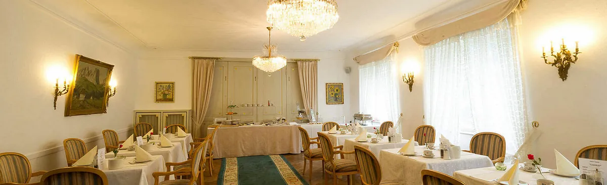 Innenansicht eines Restaurants mit mehreren Tischen, Stühlen und einem langen Buffet. Zwei große Kristalllüster hängen von der Decke. Helle Vorhänge und ein Teppich in der Mitte.